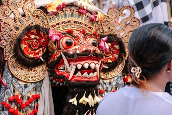 A colorful, ornate balinese mask with a person watching.